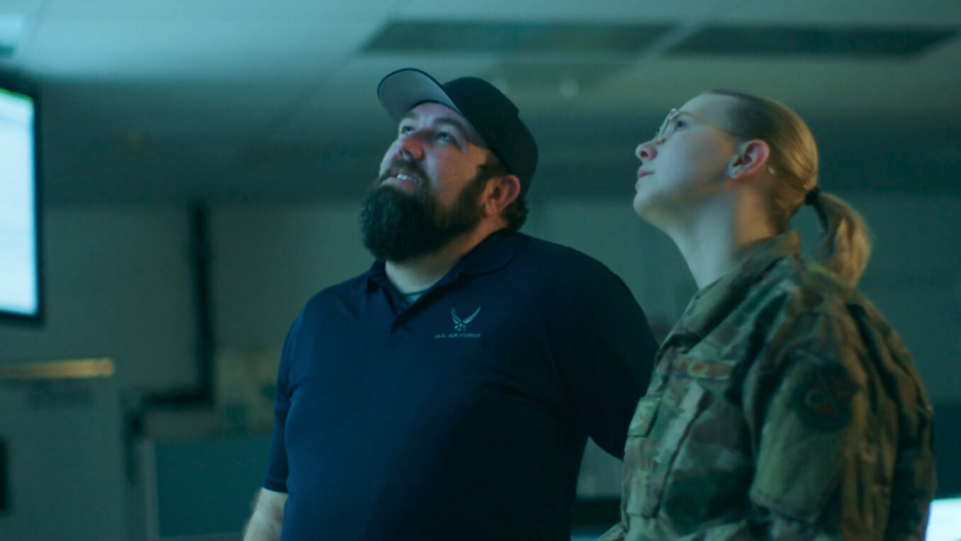 A uniformed service member and a civilian service member looking upwards towards a screen in a cyberspace operations room.